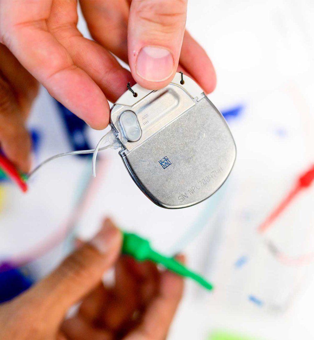 photo of a hand holding a deep brain stimulation device, which looks like a rounded, flat metallic piece with two wires coming out of it