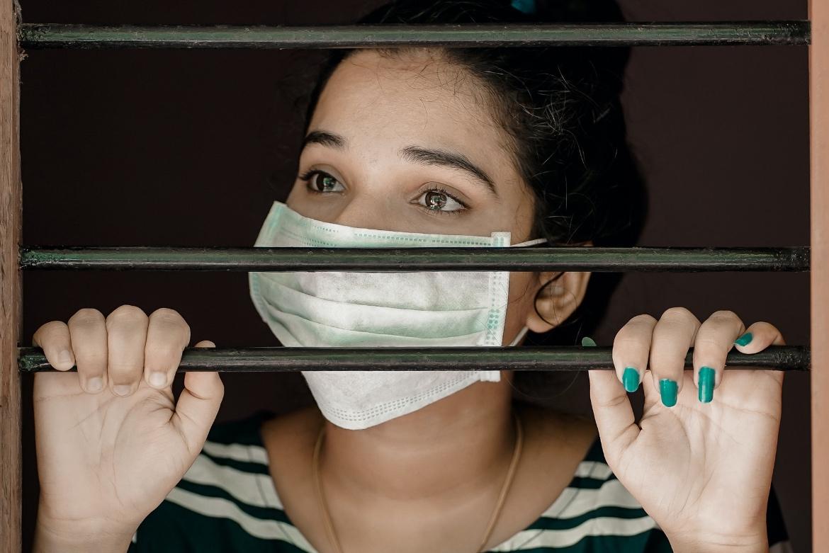 Person wearing a facial mask staring out a window with bars on it