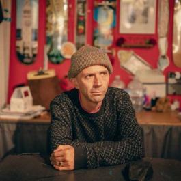 photo of a man looking to away from the camera to his left wearing a brown knitted beanie and a black knitted sweater with white zigzag pattern. In the blurred background a collection of skateboards is displayed on red walls 